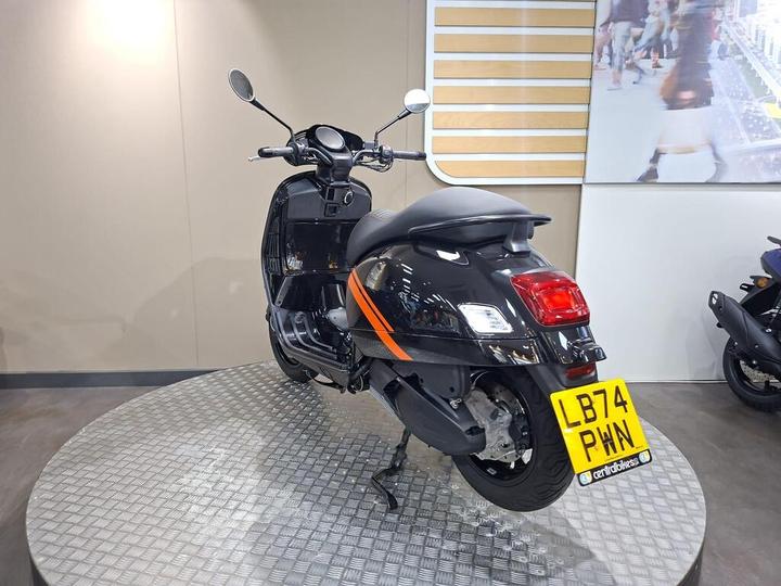 A black scooter with an orange stripe is displayed on a circular platform in a modern showroom. The rear view shows the license plate reading "LB74 PWN."