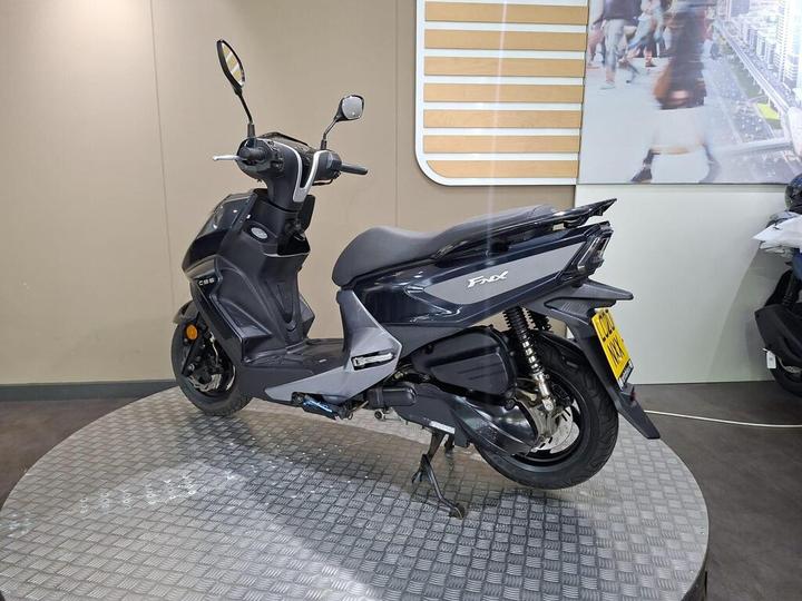 A gray scooter displayed at an angle on a circular metallic platform, with a plain background and a partial view of a cityscape in the background.