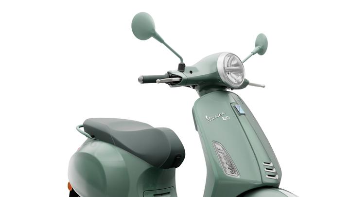 A close-up view of a mint green Vespa scooter, highlighting the front handlebar, round headlight, and side mirrors. The seat is gray, and the scooter features a metallic badge with the model name "Vespa 80."