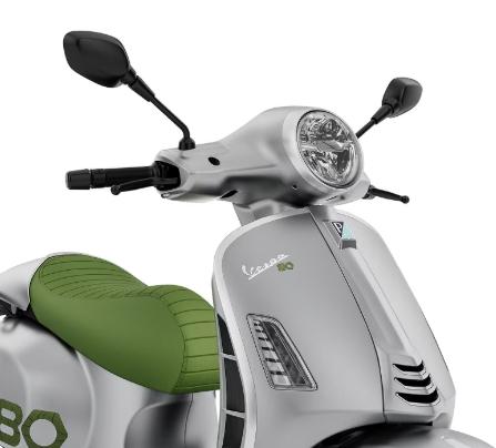 A close-up view of a silver Vespa scooter featuring a green seat, showcasing the handlebar with mirrors and the front headlight.