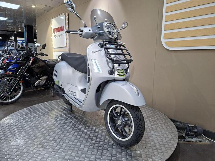 A silver Vespa scooter is displayed in a motorcycle showroom, featuring a chrome front grille, round headlight, and sporty black wheels. The background includes other motorcycles and a beige wall with wooden accents.