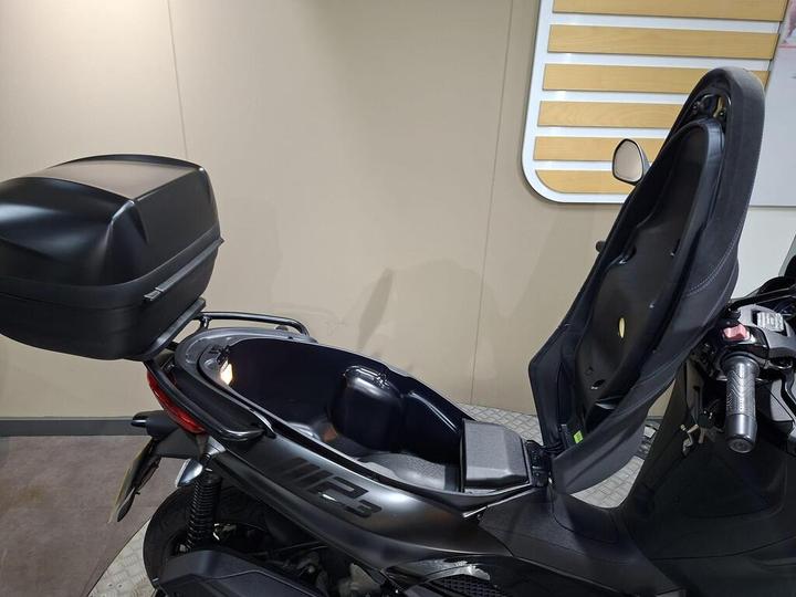 A black scooter with the seat open, revealing a storage compartment, and a mounted top box. The background features a neutral-colored wall.