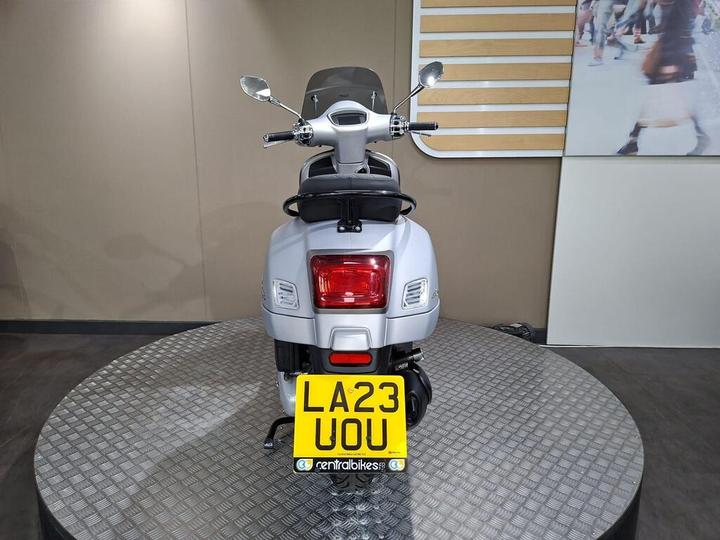 A silver scooter is displayed on a circular platform with a gray wall in the background. The rear view shows its license plate reading "LA23 UOU."
