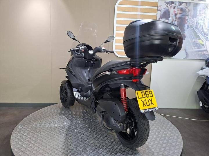 A rear view of a black three-wheeled scooter displayed on a circular platform, featuring a top case for storage and a license plate reading "LD69 XUX." The background includes a light-colored wall and promotional images.