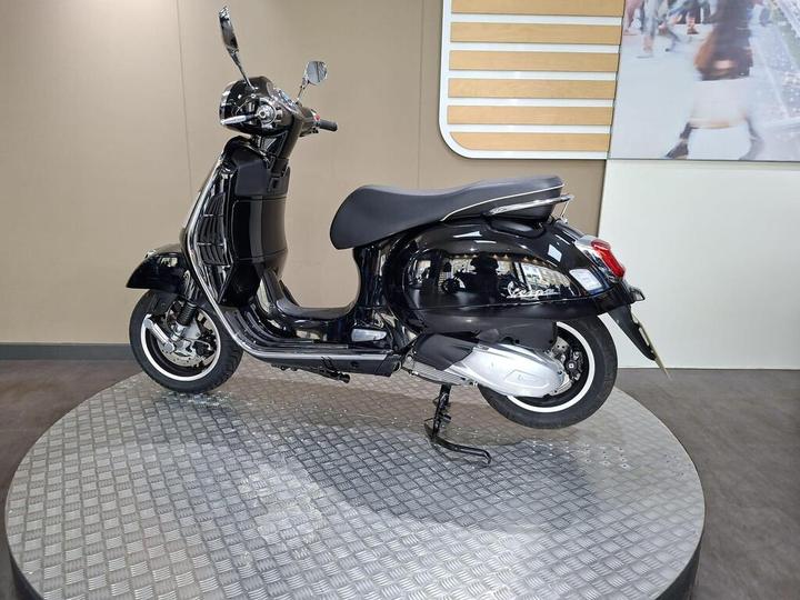 A sleek black scooter displayed on a circular platform, showcasing its design and profile, with a modern background.