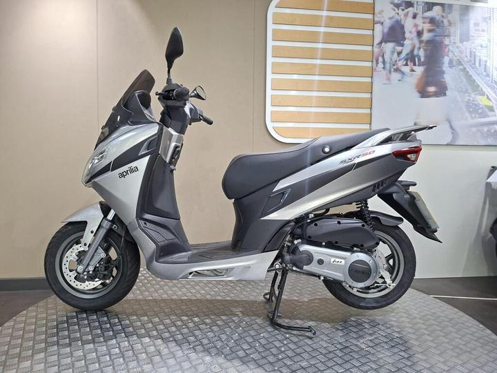 A silver Aprilia scooter parked on a textured platform against a neutral wall background. The scooter features a sleek design with a prominent windshield and a modern body style.
