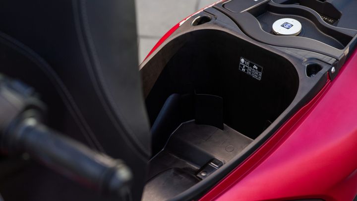 A close-up view of the storage compartment of a red scooter, showing a black interior and a circular fuel cap, with a label featuring safety instructions visible on one side.