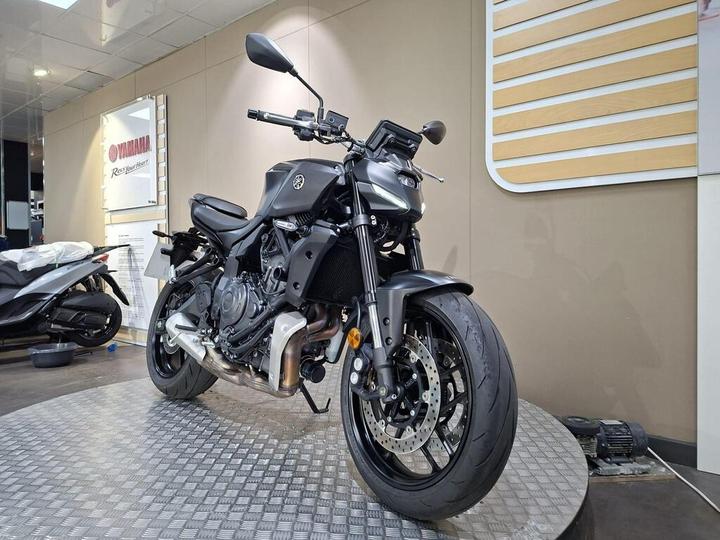 A black Yamaha motorcycle is prominently displayed in a showroom with shiny tile flooring and beige walls, alongside a partially covered lighter motorcycle in the background.