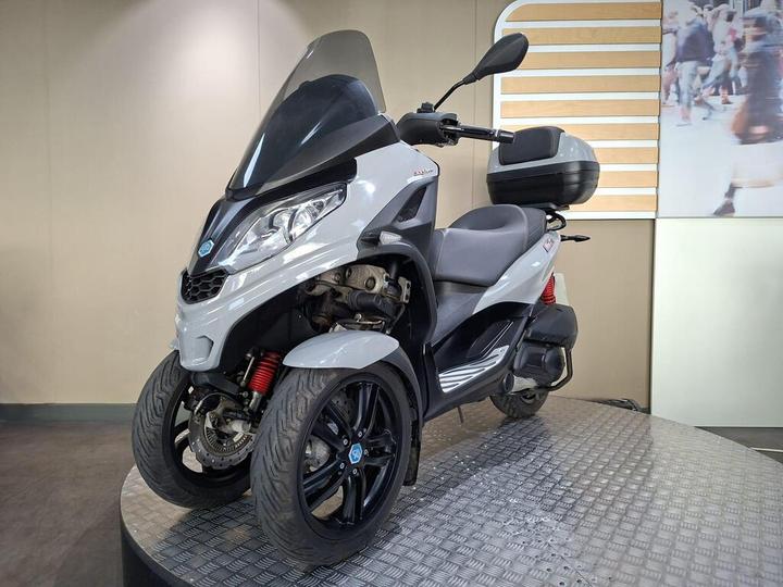 A gray three-wheeled scooter displayed on a circular platform with a textured surface, featuring a windshield, dual front wheels, and a rear storage compartment. The background includes a light-colored wall and a partially visible image of people walking.