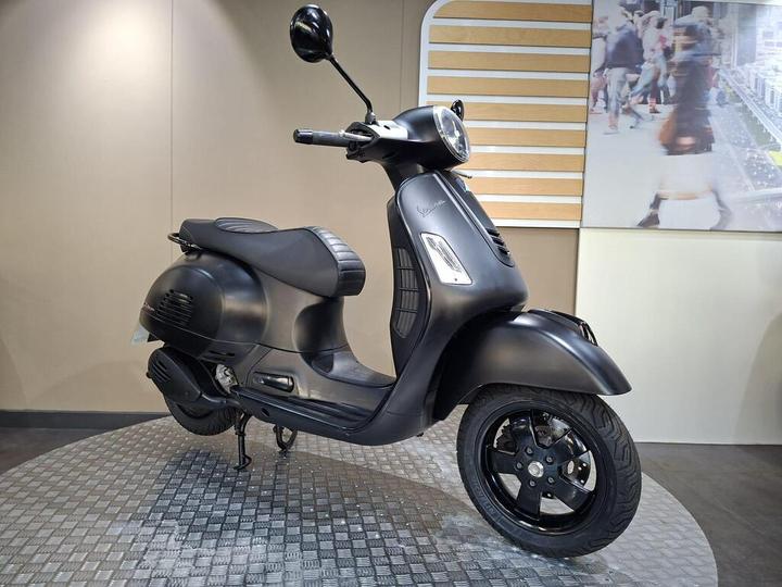 A matte black scooter displayed on a checkered floor, featuring a vintage design with a rounded headlight and a sleek, streamlined body. The background shows a light-colored wall and a blurred image of people walking.