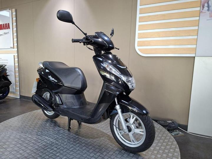A black scooter with a sleek design, positioned on a textured circular platform in a showroom, with a display board and another scooter partially visible in the background.
