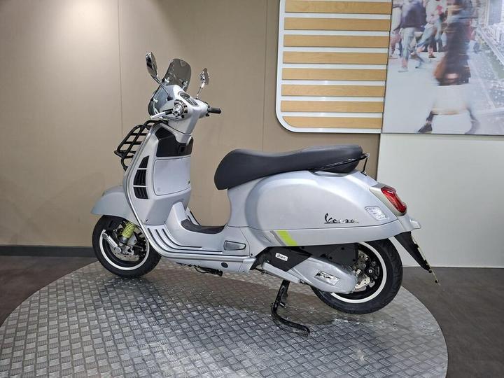 A silver Vespa scooter displayed on a circular metallic platform, set against a beige wall with a digital image of a busy city street in the background.