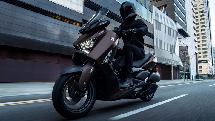 A rider in a black helmet and jacket is speeding on a brown Yamaha scooter along a city street, with modern buildings in the background.