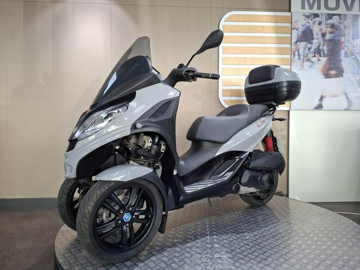 A gray three-wheeled scooter displayed on a circular platform, featuring a small windshield, saddle seat, and a rear storage compartment. The background includes a faint image of pedestrians.