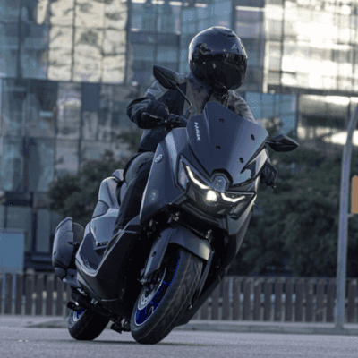 A rider in a black helmet and gear is skillfully maneuvering a sleek black scooter on an urban street, with modern glass buildings in the background.