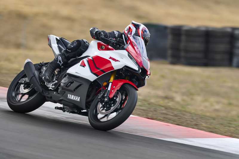 A rider leans into a turn on a red and white Yamaha motorcycle at a racetrack, with blurred tire barriers in the background.