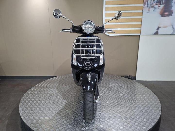 Front view of a black scooter displayed on a circular platform, showcasing its sleek design and chrome accents against a neutral background.