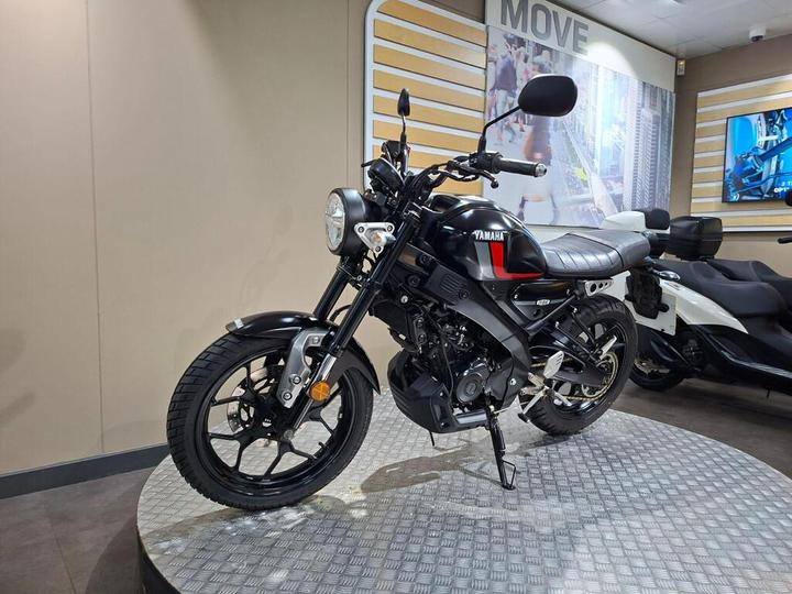 A black Yamaha motorcycle displayed on a circular pedestal in a showroom, with a modern backdrop featuring a cityscape and the word "MOVE."