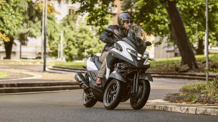 A person in a black helmet and jacket rides a three-wheeled scooter on a curved street, surrounded by trees and a green landscape.