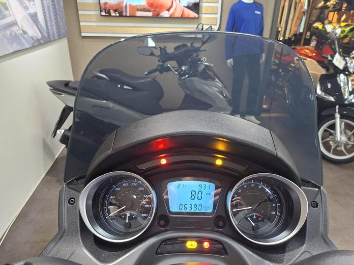 Dashboard view of a Piaggio scooter showing a digital display with speed at 80 mph, total mileage at 6390 miles, and various indicator lights. The background features motorcycles and scooters in a showroom setting.