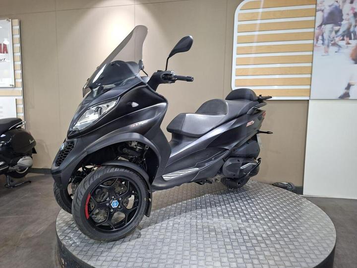 A black three-wheeled scooter displayed on a circular stand in a well-lit indoor environment, with a transparent windshield and a sleek design.