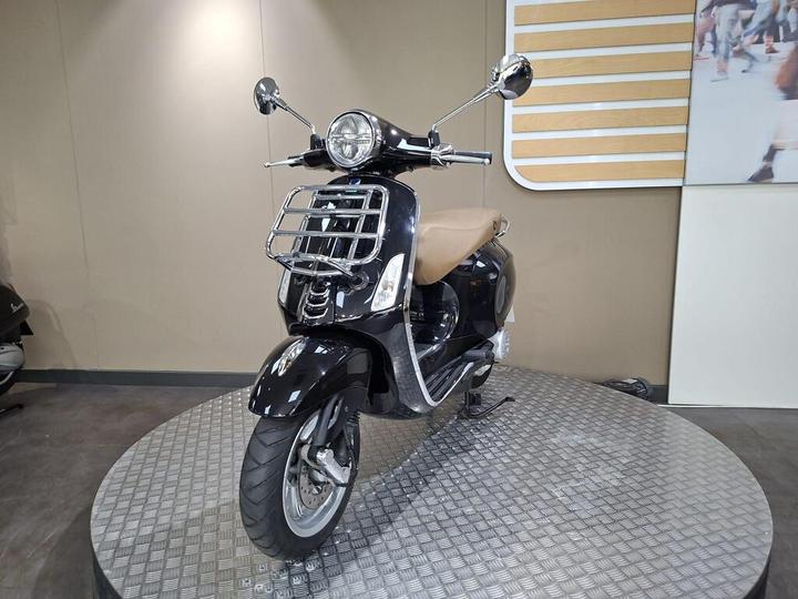 A black scooter with a chrome front rack and brown seat, positioned on a circular display platform against a neutral backdrop.