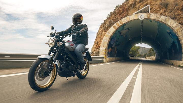 A motorcyclist rides a stylish bike on a deserted road, approaching a colorful tunnel with artistic mural decorations on its entrance, surrounded by rocky terrain and greenery.