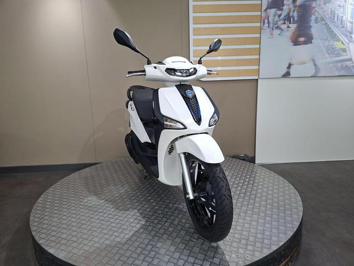 A white scooter displayed on a circular platform, featuring a sleek design with a prominent front headlight and modern styling elements. The background includes a neutral-colored wall and a digital image of a bustling cityscape.