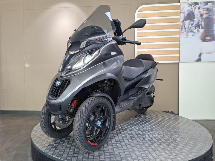 A gray three-wheeled scooter displayed on a circular platform, featuring a tinted windshield and distinct black alloy wheels. The background includes a blurred image of people walking.