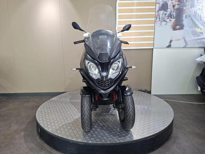 A black three-wheeled scooter displayed on a rotating platform with a gray textured surface, featuring a windshield, front lights, and prominent tires, set against a neutral background.