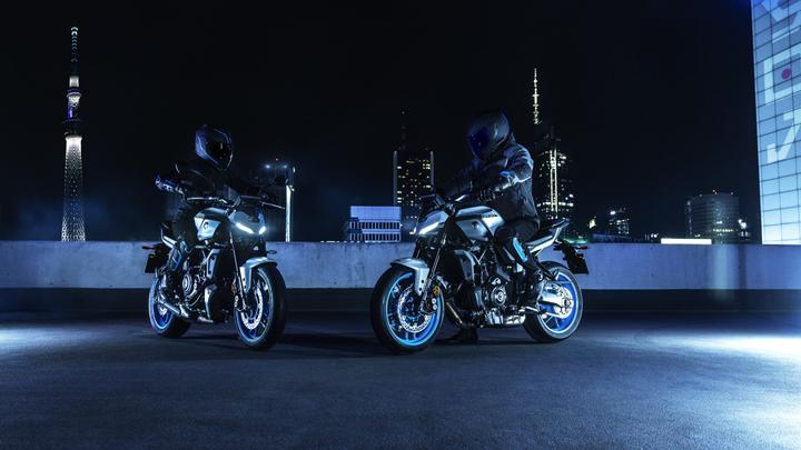 Two motorcycles are parked on a rooftop at night, illuminated by blue and white lights. Two riders, dressed in protective gear and helmets, are seated on the bikes. The background features a city skyline with tall buildings, including a lit tower.
