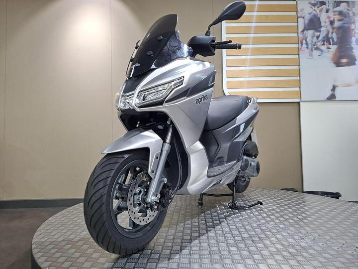 A silver Aprilia scooter displayed on a rotating platform in a modern showroom. The scooter features a sleek design with a transparent windscreen, stylish headlight, and a wide front tire. Background elements include a plain wall and a partially visible image of people in a busy urban setting.
