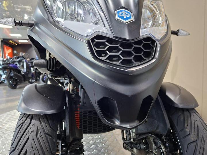 Front view of a black three-wheeled scooter with a distinct hexagonal grille, showcasing its aggressive styling and modern design elements, parked in a motorcycle showroom.