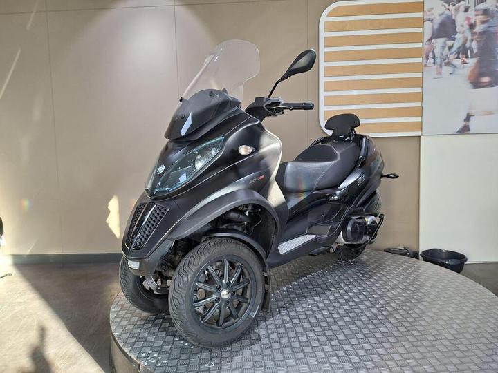 A black three-wheeled scooter with a windshield is displayed on a circular platform under soft lighting, set against a neutral wall. The scooter features a sleek design with a comfortable seat and distinctive large tires.
