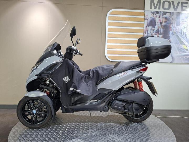 A grey three-wheeled scooter is displayed on a circular platform, partially covered with a black protective cover. It features a large windscreen, a rear storage box, and sporty black alloy wheels. The background includes a light-colored wall with a graphic of people in a busy urban setting.