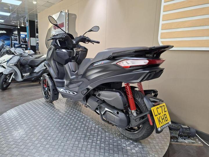 A black scooter with red shock absorbers is displayed on a checkered platform in a modern showroom, with another scooter in the background. The scooter features a sleek design and is parked beside promotional signage.