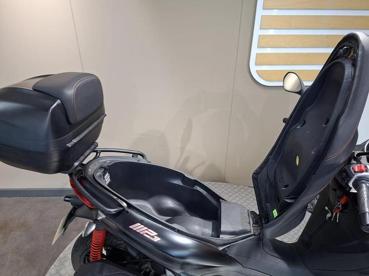 A close-up view of a motorcycle with its seat open, revealing an under-seat storage compartment. The bike features a sleek black design with a rear storage box and visible red suspension springs. The background is a neutral tone with horizontal wooden slats.
