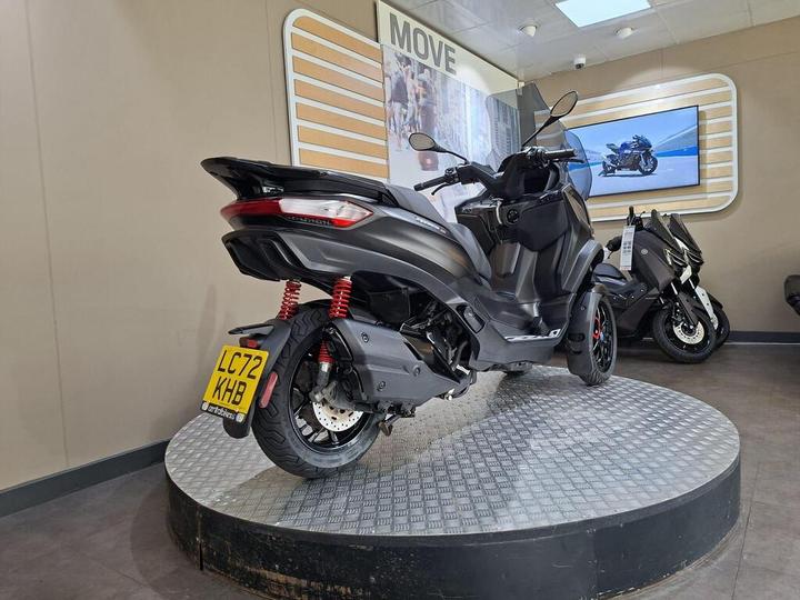 A sleek black scooter displayed on a circular platform in a showroom, featuring red suspension springs and a license plate reading "LC72 KHB." A muted background includes a monitor showcasing another motorcycle.