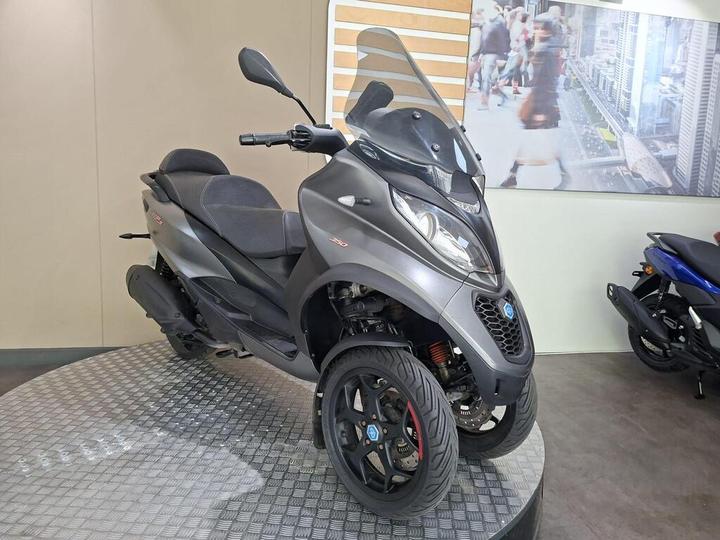 A dark gray three-wheeled scooter displayed on a circular platform in a showroom, with a modern interior and a blurred background featuring a city scene.