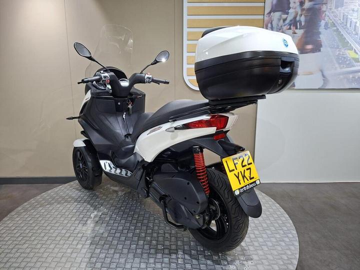 A white and black scooter with a top box, shown from the rear at an indoor display, featuring a circular floor and a background image of a busy street. The scooter displays a license plate reading "LF22 YKZ."