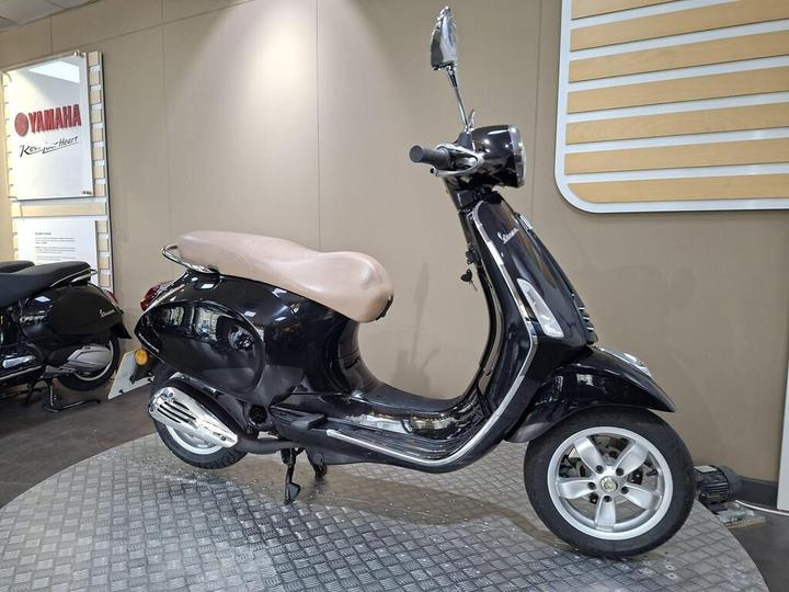 A black scooter with a beige seat is displayed in a showroom, featuring chrome details and a circular headlight, set against a neutral backdrop with Yamaha branding and promotional materials.