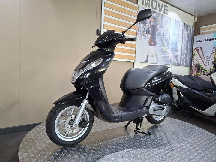 A black scooter is displayed on a circular stand with a textured surface, set against a neutral background featuring promotional images and text related to urban mobility.