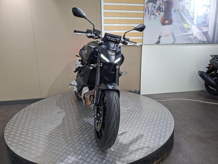 A black Yamaha motorcycle is displayed on a circular platform with a textured surface, facing forward. The background features a light-colored wall and a digital screen displaying a city scene.