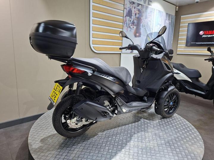 A black three-wheeled scooter displayed on a circular platform in a showroom, featuring a rear storage compartment and a sleek design, with a beige wall and Yamaha branding in the background.