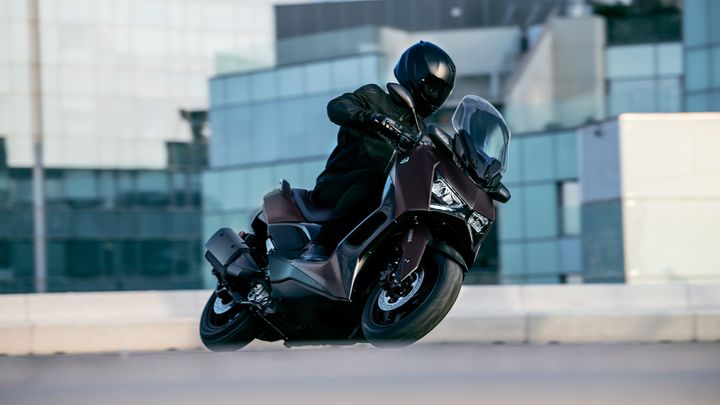 A rider in a black outfit and helmet leans into a turn on a dark motorcycle against a backdrop of modern glass buildings.