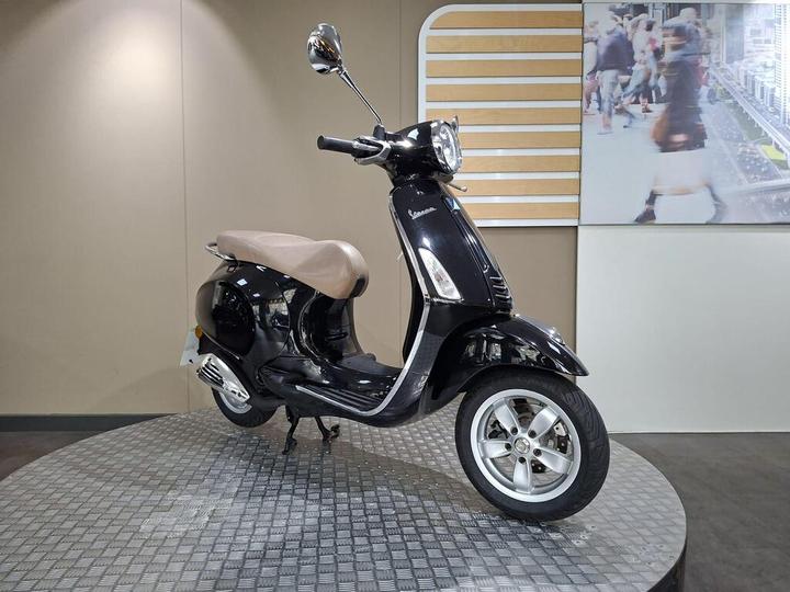 A black scooter with a tan seat is displayed on a circular platform, featuring chrome accents and a classic design. In the background, blurred pedestrians are visible on a large screen.