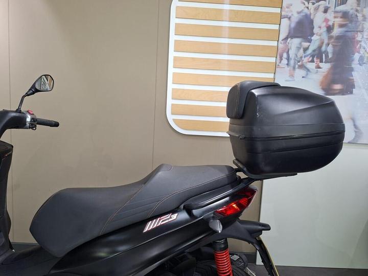A black scooter with a large rear storage box is displayed against a neutral backdrop, featuring a design of horizontal wooden slats and a blurred crowd scene.