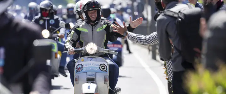 A group of motorcyclists and scooter riders participate in a parade, with one rider on a scooter smiling and waving to the crowd while others ride behind. The scene is lively, showcasing camaraderie and enthusiasm among the participants.