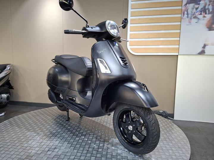 A sleek, matte black scooter is displayed on a circular platform against a neutral background, featuring a modern design with a prominent headlamp and textured seat.