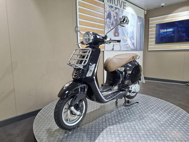 A black scooter with a vintage design is displayed on a circular, textured platform against a backdrop of light-colored walls and digital screens. The scooter features a brown seat and chrome accents, showcasing a modern yet classic aesthetic.